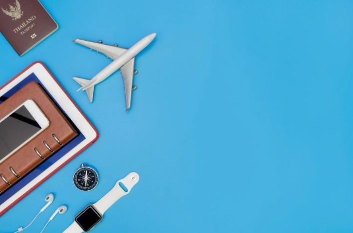 A miniature plane, passport, tablet, smartwatch, compass, headphones, cell phone, and notebook are arranged on a bright blue background.